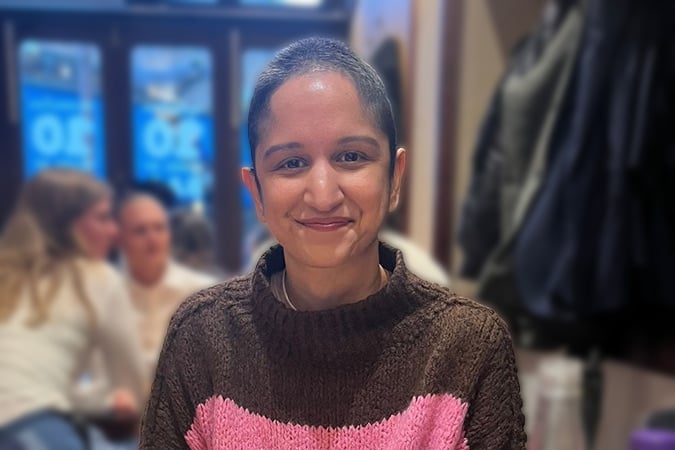 A person with short hair and a brown and pink jumper, fresh from a recent breast cancer scare, smiles at the camera in a cosy indoor setting, with other people blurred in the background.