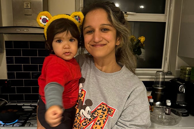 A smiling woman stands in a kitchen holding a baby wearing a bear-ear headband and red shirt. The woman wears a grey Mickey Mouse sweatshirt.