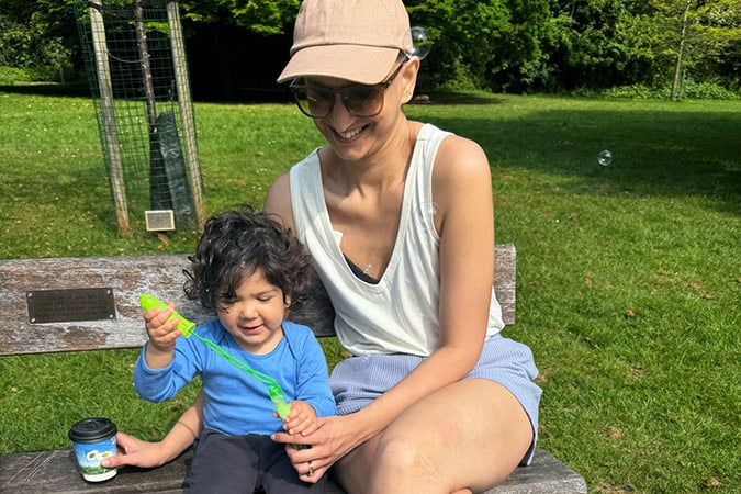 A smiling woman in sunglasses and a cap sits on a park bench with a young child blowing bubbles, both enjoying a sunny day. The child holds a bubble wand and a small cup. Green grass and trees are in the background.