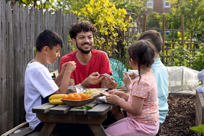 A man and three children sit at a picnic table in a garden, eating and talking together. Surrounded by plants and trees, they enjoy bananas and oranges on the table—perfect examples of healthy picnic ideas for families to share outdoors.
