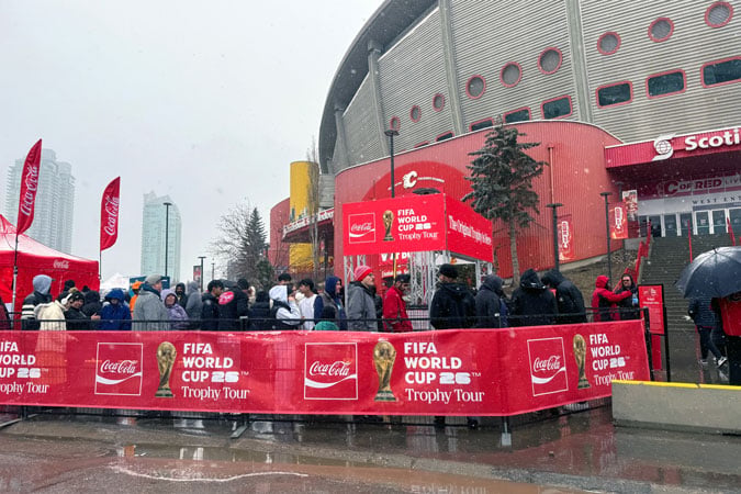 People queue outside a stadium in winter weather for the FIFA World Cup Trophy Tour, surrounded by Coca-Cola banners and red fencing—a striking example of health-harming sports sponsorship on display.
