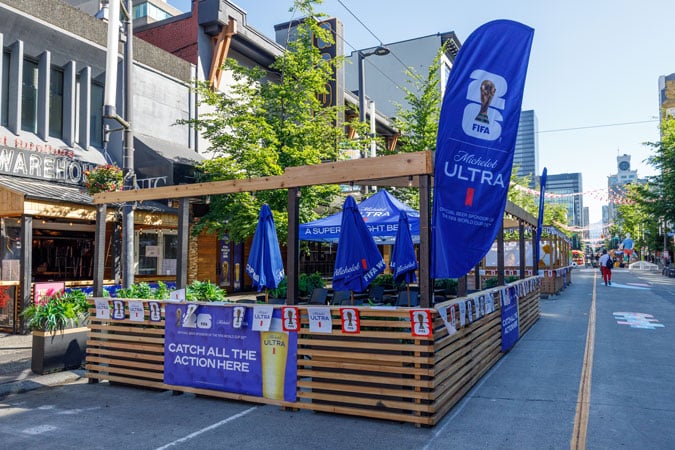 A street pavement café with branded banners and flags, outdoor seating, and parasols, set up beside restaurants on a sunny day. The decorations promote a sporting event and a beverage brand through health-harming sports sponsorship.