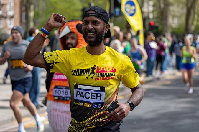 A smiling runner in a yellow London Landmarks Half Marathon 2027 shirt with a PACER bib raises his fist in celebration during a race, with other runners and spectators visible in the background.
