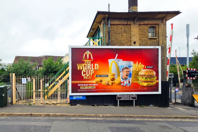 A billboard advertises McDonald's World Cup Meal, showing chips, a burger, a drink, and a footballer, highlighting the issue of health-harming sports sponsorship. The sign is next to a level crossing and an old brick building, surrounded by fences and greenery.