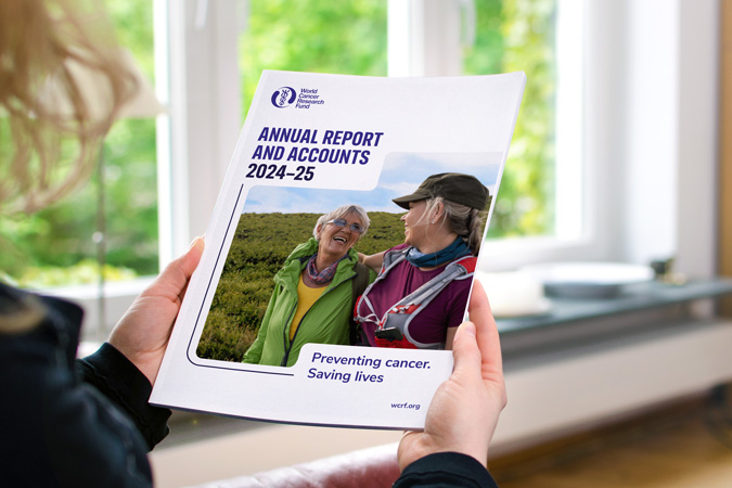 A person holds the WCRF Annual Report 2024–25, featuring two smiling women outdoors, with the text Preventing cancer. Saving lives on the cover.