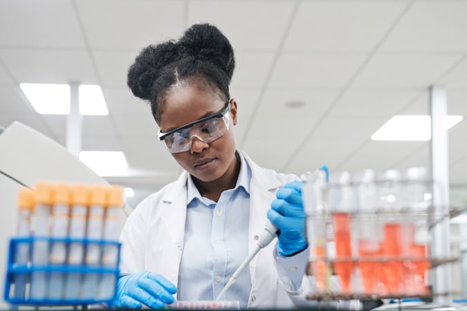A scientist wearing safety glasses and gloves uses a pipette in a lab, working with test tubes filled with various coloured liquids—an example of how our research impact advances scientific discovery.