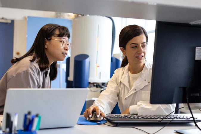 Two women in a laboratory, one wearing a white coat, sit at a desk working together on a computer, reviewing data for the WCRF Annual Report 2024–25. A laptop, pens, and scientific equipment are visible in the background.
