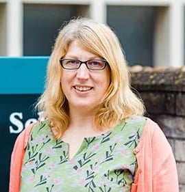 Dr Sarah Lewis, a woman with blonde hair and glasses, stands smiling near a sign that reads Bristol Medical School. She is wearing a green patterned top and a light coral cardigan. The background includes a building and windows.