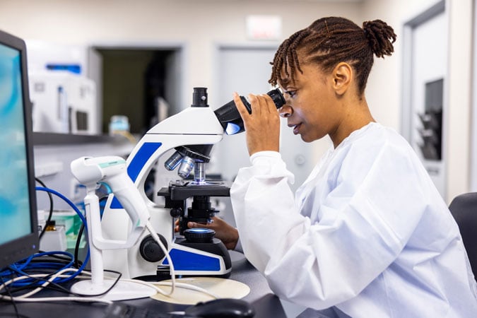 A scientist wearing a white lab coat looks into a microscope at a laboratory workstation, with laboratory equipment and a computer also visible nearby.