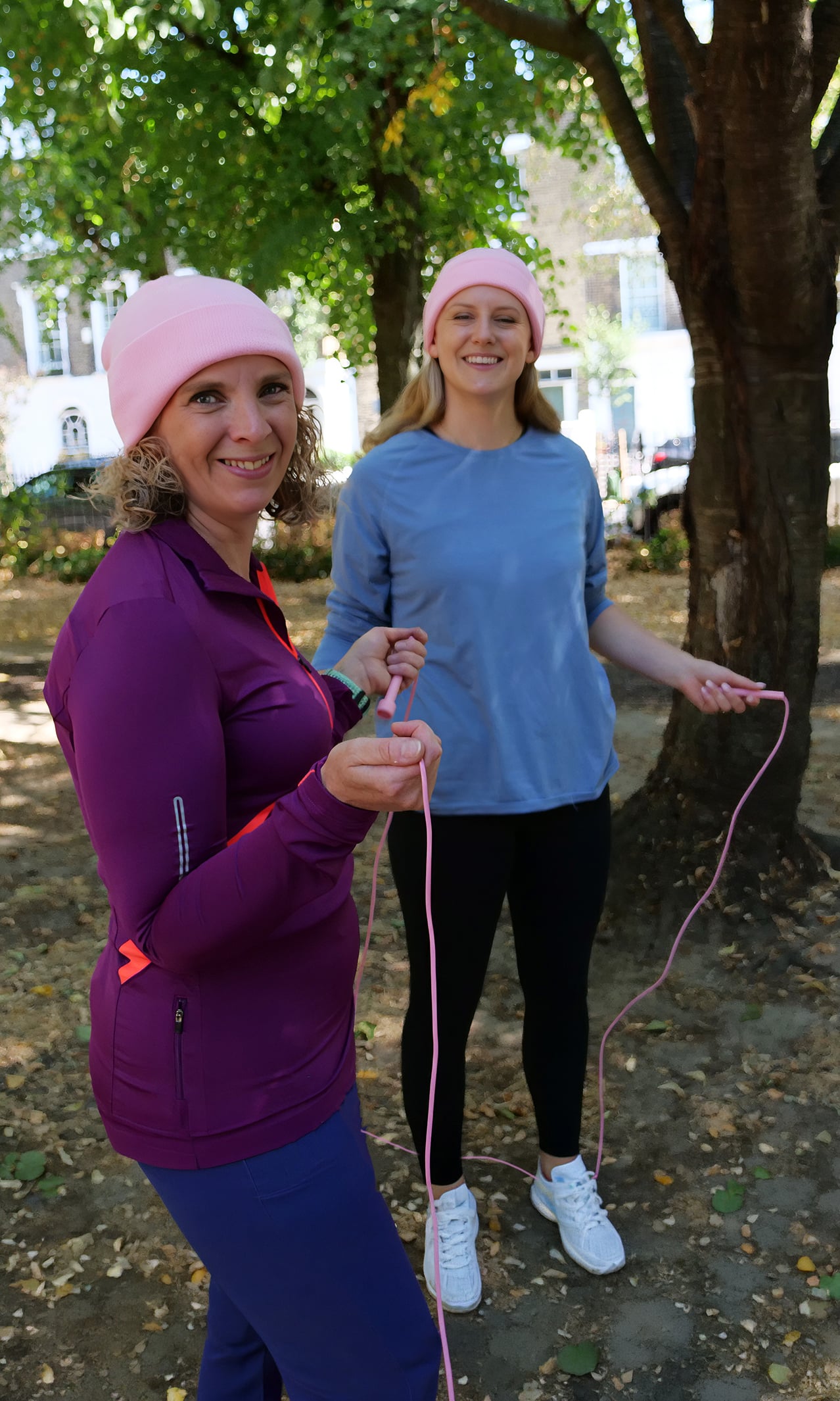 Two women in pink hats are outdoors under trees, smiling and holding a skipping rope together as they support the World Cancer Research Fund. One wears a purple jacket, the other a blue shirt. It’s a sunny day with leaves scattered on the ground.