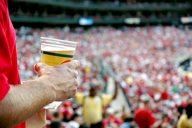 A person holding a plastic cup of beer stands in front of a crowded stadium with many people in red shirts, suggesting a lively sports event atmosphere—highlighting the prevalence of health-harming products sport sponsorship at such popular gatherings.