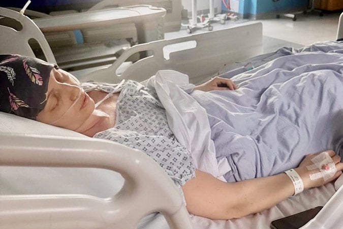 A woman wearing a headscarf lies in a hospital bed, eyes closed, with a hospital gown and a drip in her hand.