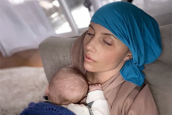 A woman wearing a blue headscarf rests on a sofa with her eyes closed, holding a baby who is snuggled against her chest.