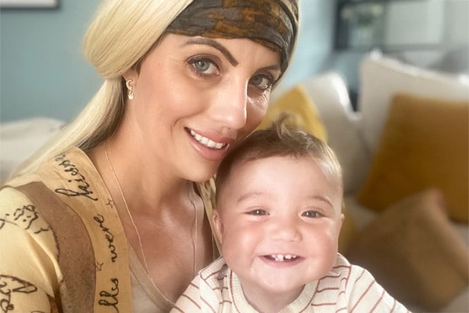 A woman with a headscarf smiles whilst holding a happy baby with short brown hair, both sitting on a sofa with yellow cushions in the background.