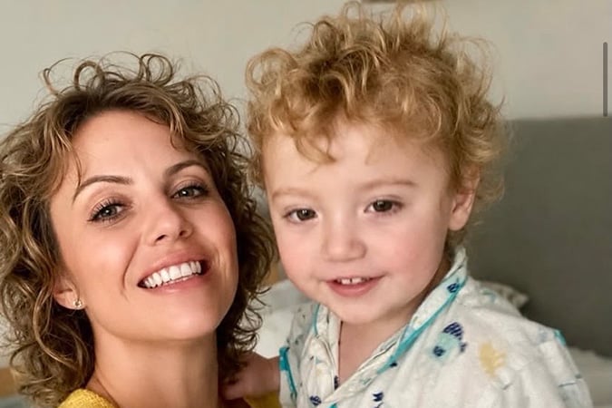 A woman with curly hair smiles next to a young child with curly blond hair, both looking at the camera in a cosy indoor setting.
