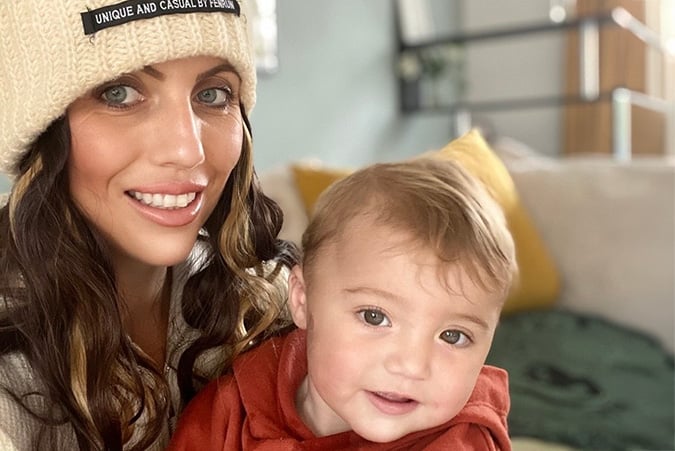 A woman wearing a knitted hat smiles next to a toddler in a red jumper. They are sitting on a sofa with yellow and green cushions in the background.