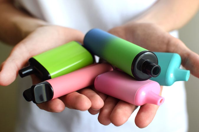 A person holds several colourful disposable vape pens, including green, blue, pink, and black, in their hands—evoking concerns similar to those raised by health-harming sports sponsorship that can glamourise risky behaviours.