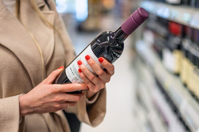 A person with red nail varnish holds a bottle of red wine with a purple cap whilst shopping in a shop aisle, pausing to read the cancer prevention alcohol labels displayed on the bottle.