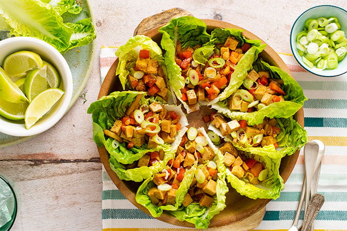 A wooden bowl with lettuce wraps filled with diced tofu—a delicious way to explore the relationship between soya consumption and breast density—red peppers, and sliced spring onions, surrounded by lime wedges and extra spring onions on a striped cloth.