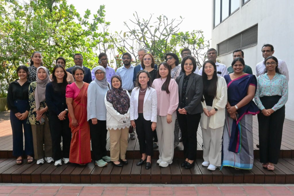 A group of 21 adults stands together outdoors on wooden steps, smiling at the camera. The group, gathered for a discussion on benchmarking policies, includes men and women in a mix of formal and casual attire, with greenery and a building in the background.