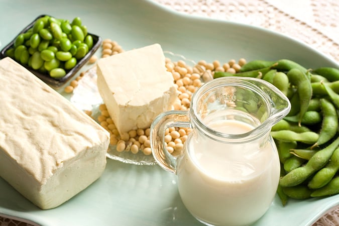 A plate with tofu blocks, a glass jug of soya milk, soya beans, and edamame pods and beans, all displayed on a light tray.