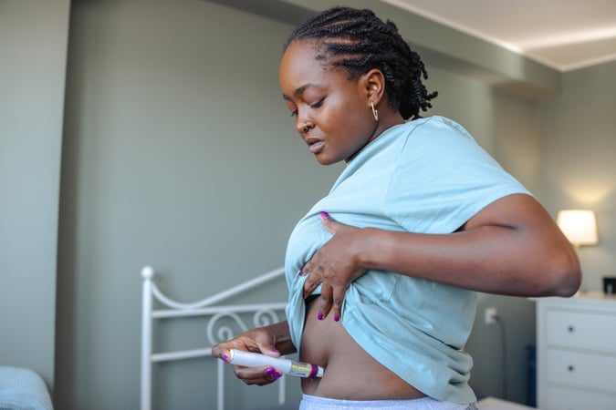 A person lifts their shirt and injects GLP-1 medication into their abdomen while standing in a bedroom, a routine that has sparked recent discussions about the possible links between GLP-1 medications and cancer.