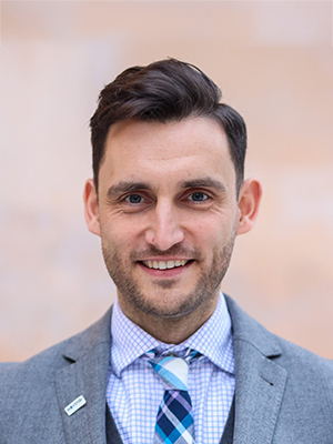 Maik Dunnbier, a man with short dark hair and beard, wearing a grey suit, patterned tie, and checked shirt, smiling in front of a blurred light background.