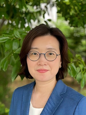 Professor Jin-Kyoung Oh, a woman with short brown hair and glasses wearing a blue blazer and standing outdoors in front of green foliage.