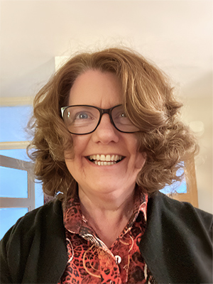 Sheila Gilheany, a woman with curly light brown hair and glasses smiling broadly. She is wearing a patterned red shirt and a dark jacket. The background is softly lit indoors with a window and light-coloured walls.