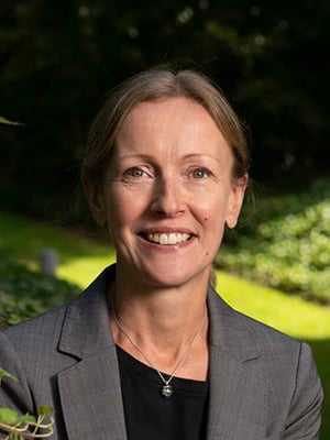 Susan Taylor, a woman with light brown hair, tied back, smiling while wearing a grey blazer and silver necklace, standing outdoors with greenery in the background.