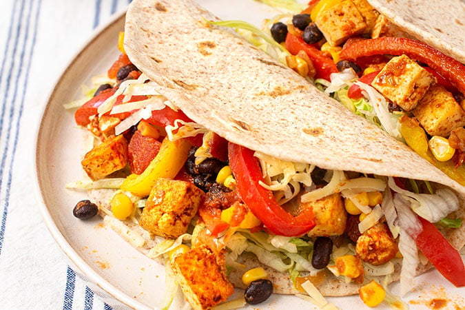Two soft tortillas filled with spiced tofu cubes—highlighting the benefits of soya consumption and breast density—black beans, sweetcorn, shredded lettuce, red and yellow peppers, and diced tomatoes, served on a white plate.