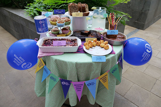 A table with assorted baked goods, muffins, brownies, drinks, and blue balloons. Colourful bunting hangs from the front, reading Preventing Cancer Saves Lives. The scene appears festive and inviting.