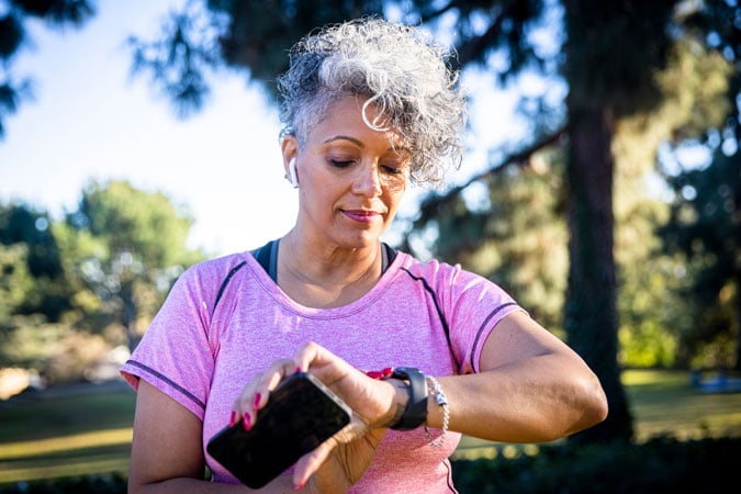 A woman with short curly grey hair wearing a pink shirt looks at her smartwatch outdoors, holding a mobile phone in one hand. Trees and blue sky are in the background.