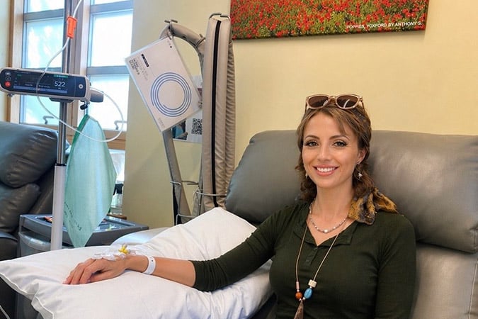 A woman sits in a medical chair, smiling with a cannula in her arm—a moment of strength as she faces breast cancer during pregnancy. She wears a green top and necklaces, sunglasses on her head, and a floral painting hangs on the wall behind her.