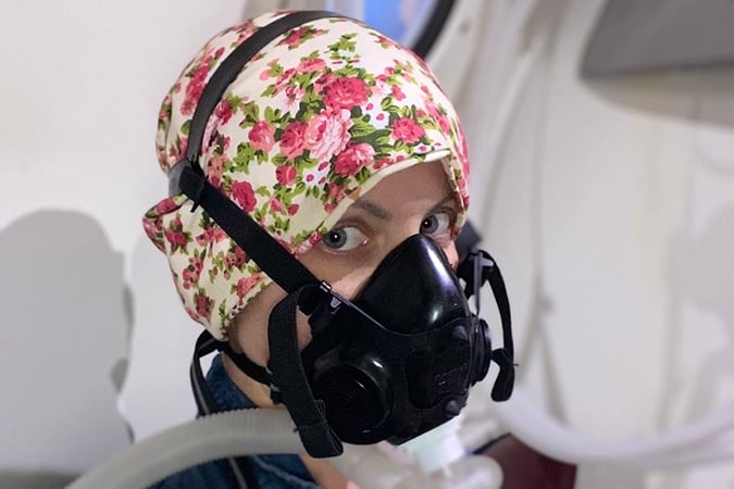 A person wearing a floral headscarf and a black medical mask is looking at the camera inside what appears to be a medical or laboratory setting, possibly reflecting the experience of individuals facing breast cancer during pregnancy.