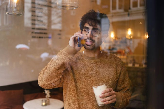 A young person wearing a brown jumper and glasses talks on a mobile phone while holding a glass of iced coffee, seen through a window.