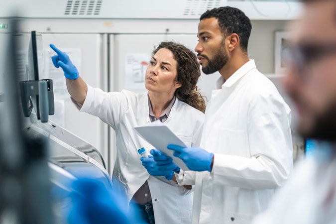 Two young scientists in lab coats and gloves examine cancer research data on a computer screen.