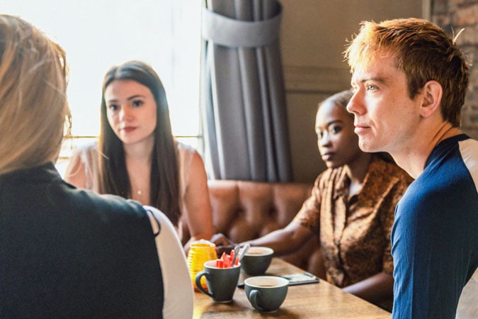 Four people sit around a wooden table in a café, talking and drinking coffee.